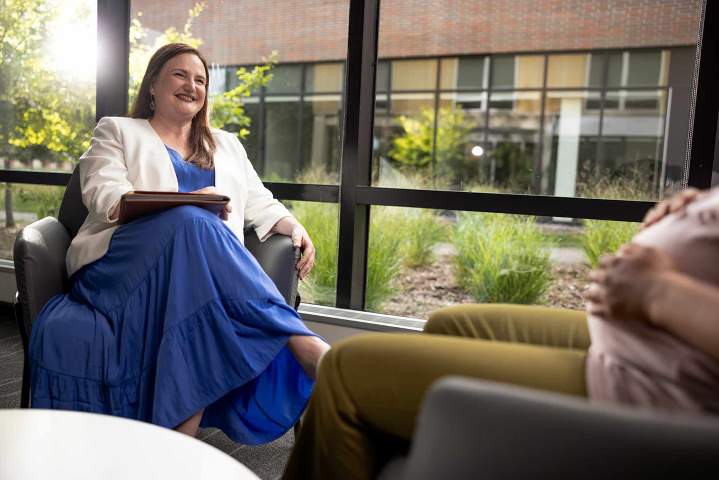 An individual seated in a chair indoors, holding a book and smiling during a conversation, with large windows overlooking greenery in the background.