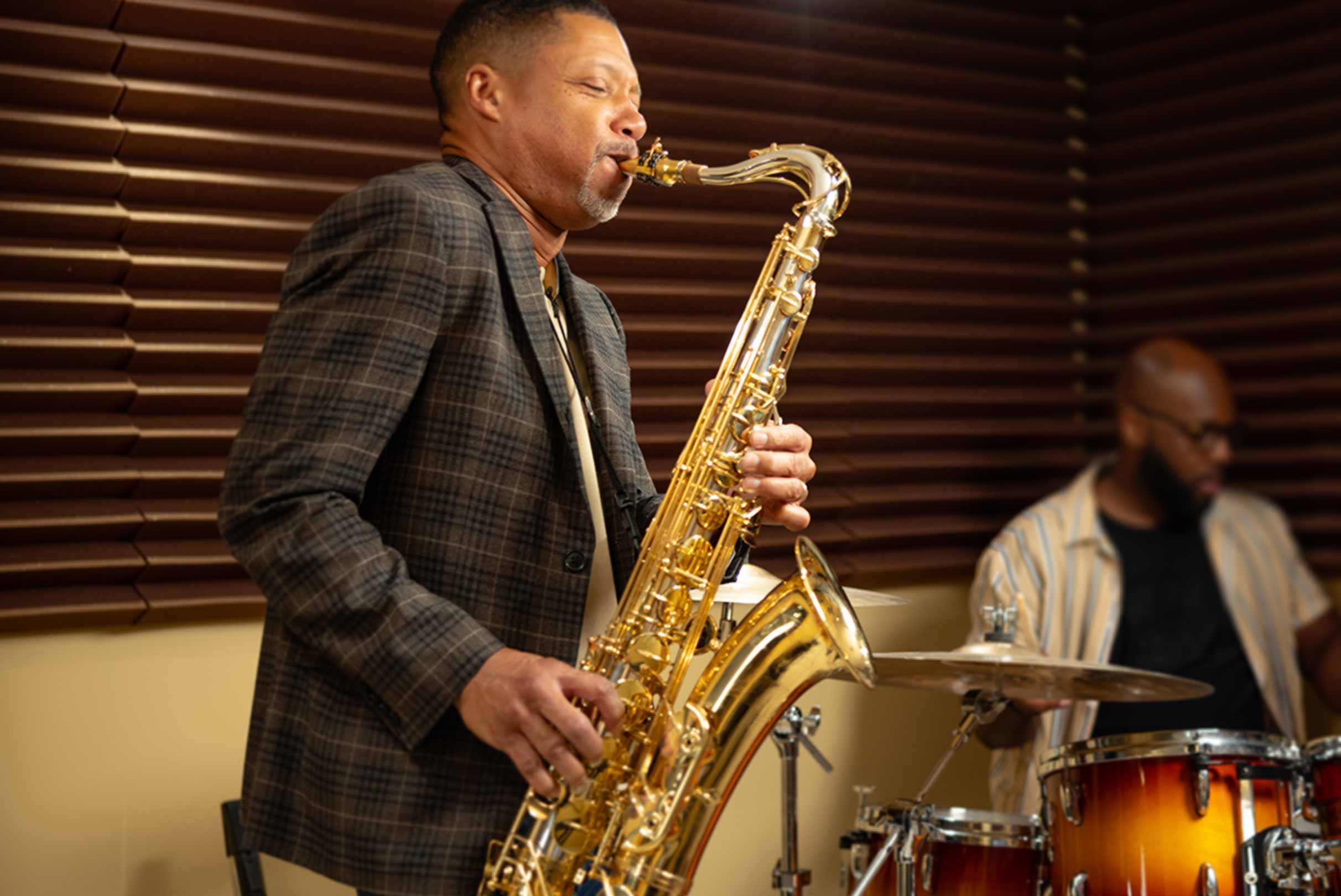  A saxophonist in a plaid jacket playing his instrument with fervor, accompanied by an out-of-focus drummer in the background, in a room with wooden paneling.