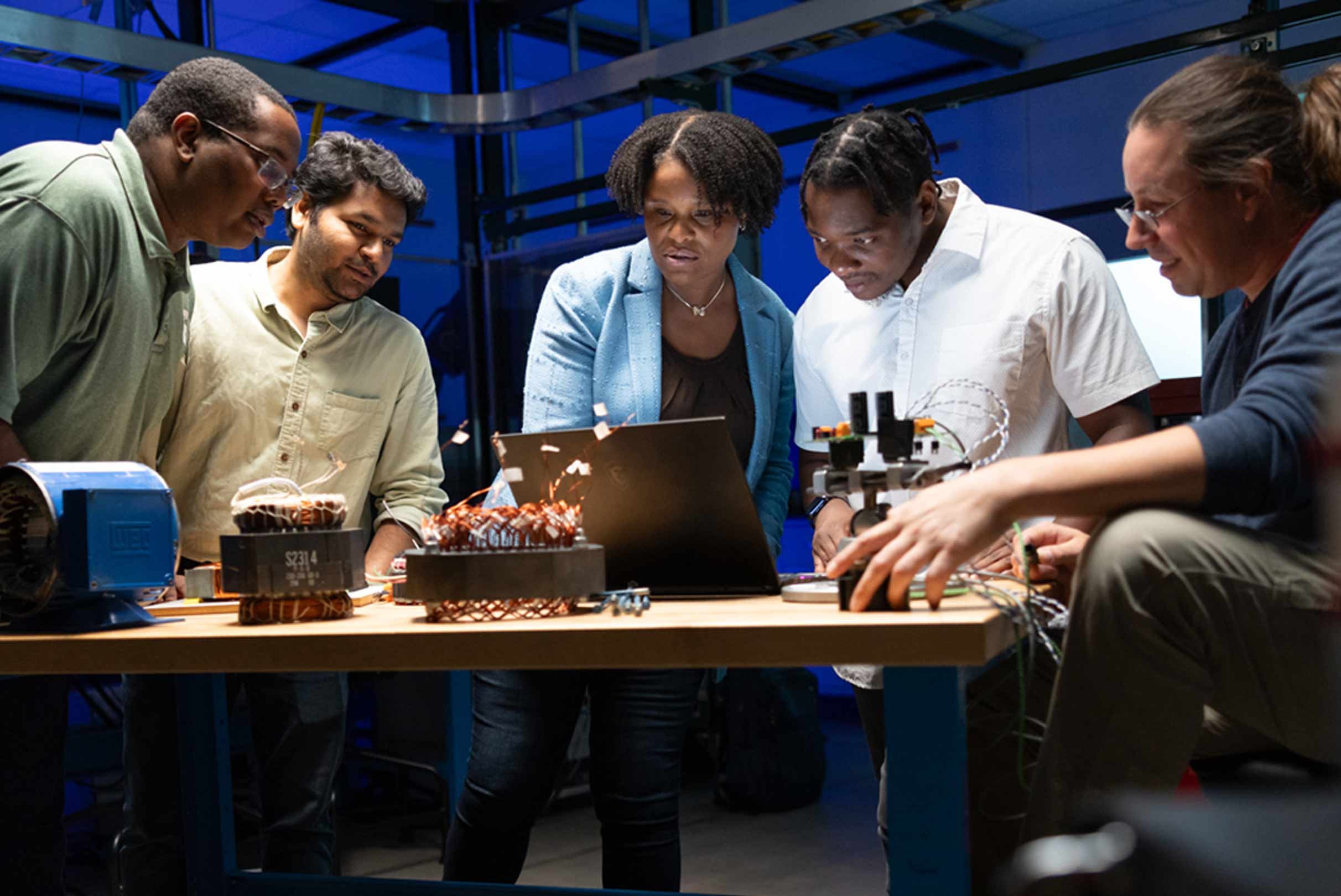 A diverse team of five collaborating on an engineering project in a tech-laden workshop, with one person operating a laptop and others examining electronic components.