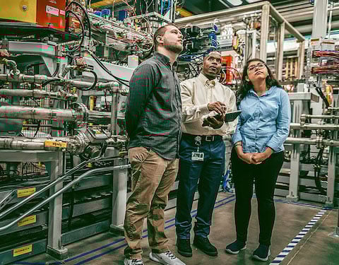 A faculty member holding a clipboard chats with students in the Facility for Rare Isotope Beams