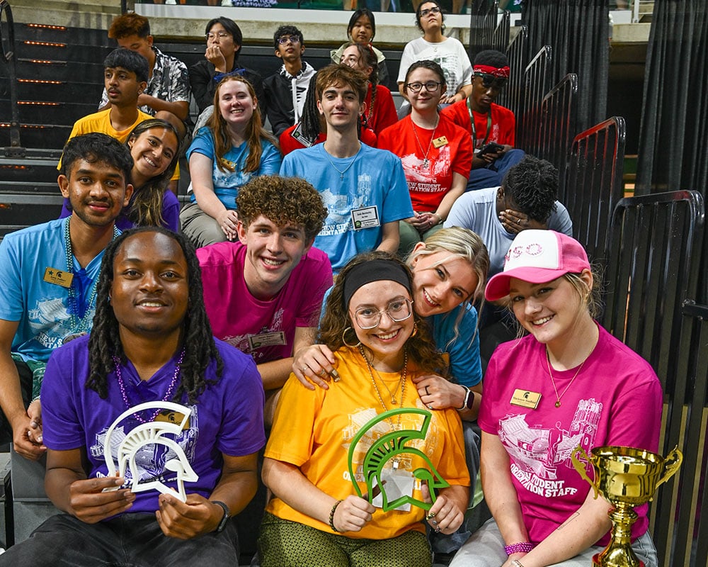 A group of students sit on bleachers. They are all wearing different colored shirts and smiling at the camera. The students in the front are holding two 3D printed Spartan Helmets, one green and one white.