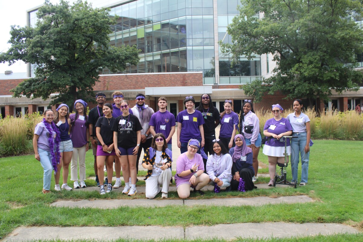 A group of students stands together in a grassy area in front of a large, windowed building. They are looking at the camera and smiling and predominantly wearing purple.
