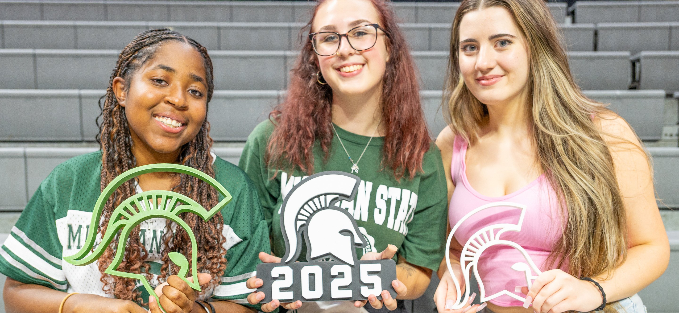 Three students sit in a row on a set of bleachers. They are looking at the camera and smiling. They are all holding a large spartan helmet in their hands, with the center students reading "2025"