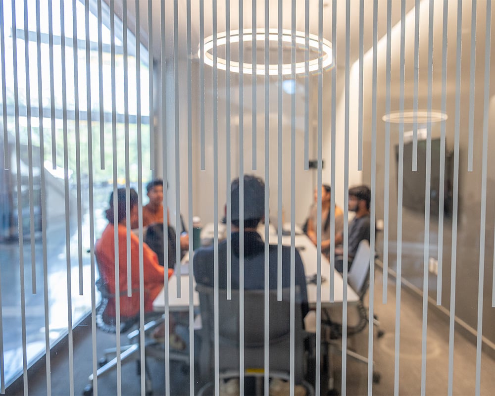 Students sitting with their backs to the camera around a long table. The picture is looking through a glass window with faint lines running down it, foregrounding the group at the table.
