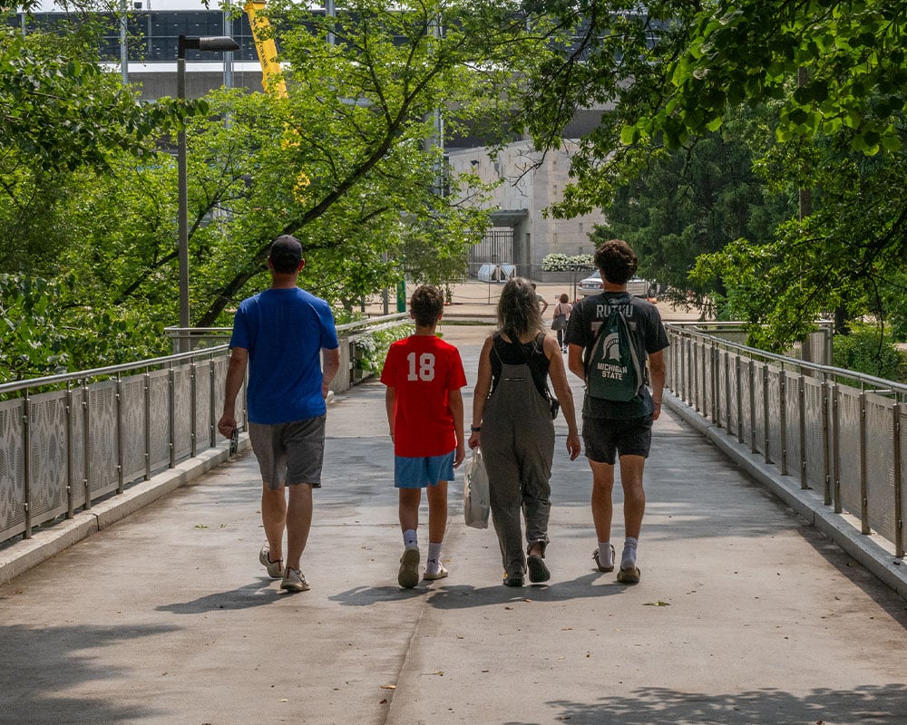 Four people standing side by side are walking away from the camera down a bridge surrounded by green foliage. 