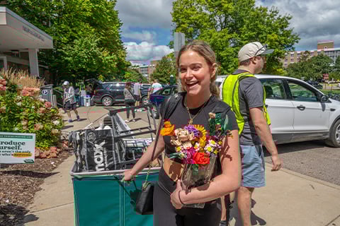 A student smiles while looking at the camera while pulling a large green tote full of living supplies. The students other hand is full of additional items. 