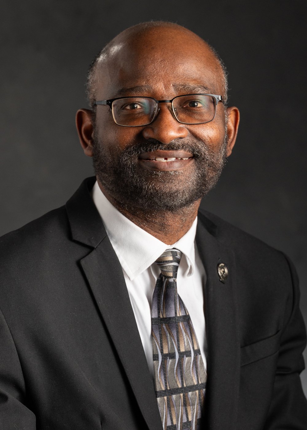Headshot of Titus Awokuse, Vice Provost Dean of International Studies and Programs, smiling and wearing a black suit, white shirt, and tie, standing against a dark gray background.