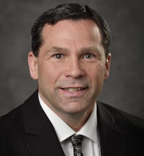 Headshot of Ryan Thelen, smiling, wearing a black suit and white shirt, standing in front of a gray background.