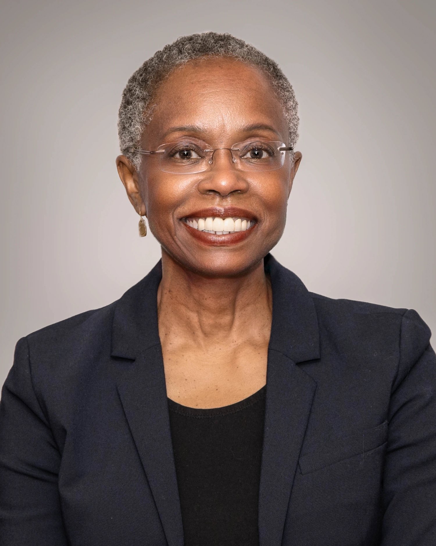 A headshot of Teresa Mastin, smiling and wearing a black business suit, standing against a gray background.