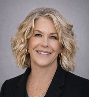 Headshot of Sue Rankin, smiling, wearing a dark blazer and green blouse against a gray background.