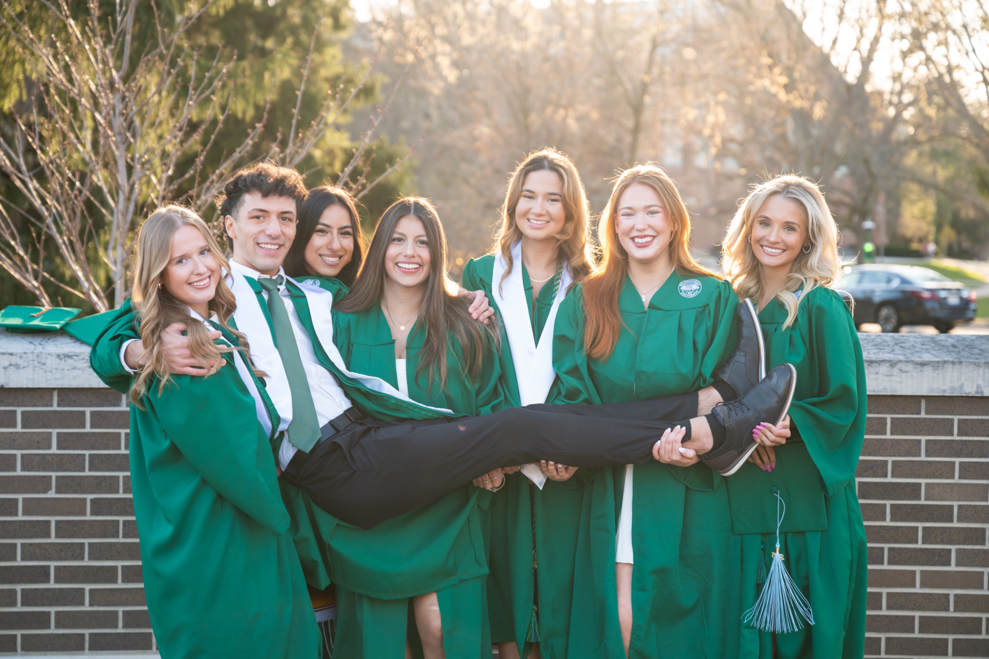 Seven undergrads in graduation regalia pose for a picture in front of a brick wall. 