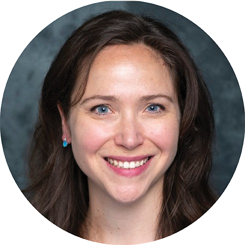 Portrait in circle frame. Subject viewed from the shoulders up, and is looking at the camera smiling with long brown hair against a blue grey background.