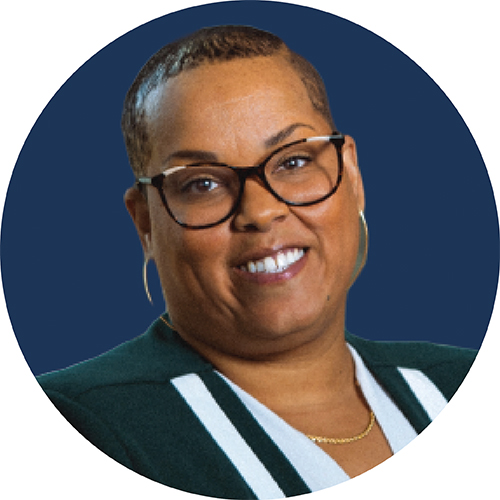 Portrait in a circle frame. Subject is seen from the shoulders up wearing dark glasses and smiling at the camera. Their hair is cut short and they are against a blue background.