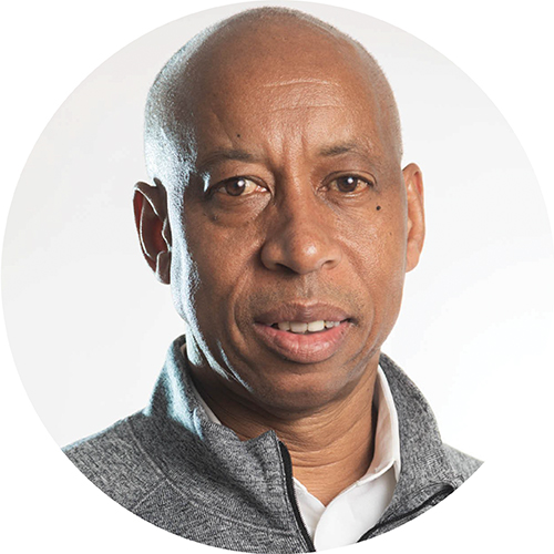 Portrait in a circle frame. Subject is looking at the camera and can be seen from the shoulders up. They are bald and standing against a white background wearing a grey collared shirt.
