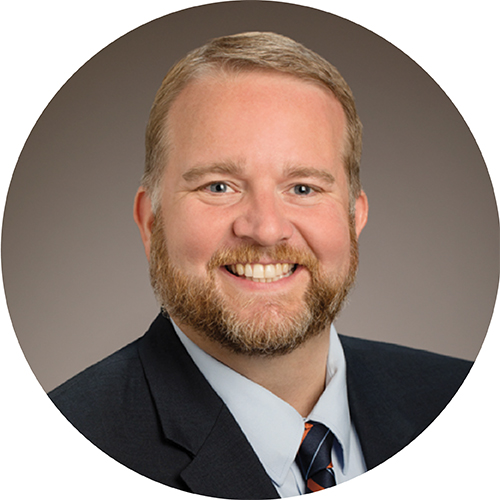 Portrait framed in a circle. Subject is looking at the camera and smiling, seen from the shoulders up. They are wearing a suit and tie, with short sandy blonde hair, standing in front of a tan backdrop.