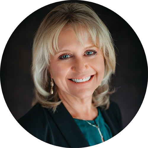 Portrait in a circle frame. Subject is seen from the shoulders up. They are smiling while looking at the camera, with blonde hair to their shoulders while wearing a dark blazer with a teal shirt against a dark backdrop.