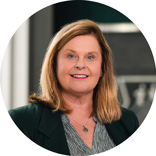 Portrait framed in a circle. Subject is looking at the camera and can be seen from the shoulders up. They are standing in an open space, wearing a dark jacket and blouse, with blonde hair to their shoulders.