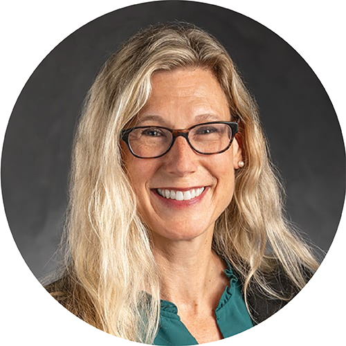 Portrait framed in a circle. Subject is looking at the camera and smiling, seen from the shoulders up. They are wearing a dark jacket and green shirt, wearing glasses, with blonde hair past their shoulders, standing against a dark grey background.