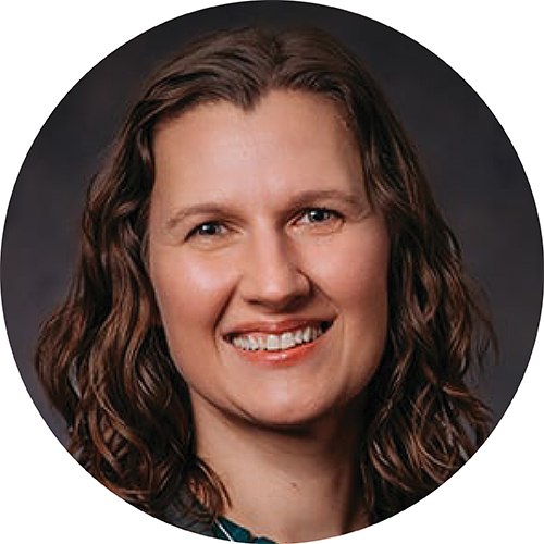 Portrait in a circle frame. Subject is smiling while looking at the camera, seen from the shoulders up. Their hair is brown and down to their shoulders and they are standing against a dark background.