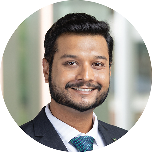 Portrait in a circle frame. Subject is looking at the camera and smiling. They are seen from the shoulders up, and are wearing a suit jacket with a white shirt a blue-green tie. Their hair is short and dark and they have a beard. They are standing in a building with the background blurred.
