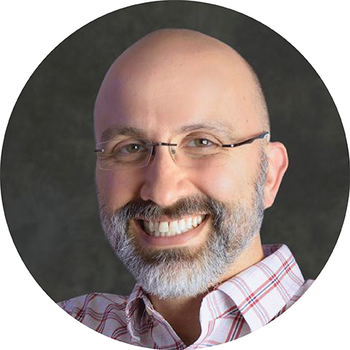 Portrait in a circle frame. Subject is looking at the camera and smiling, seen from the shoulders up. They are wearing glasses and a plaid shirt, against a grey background.