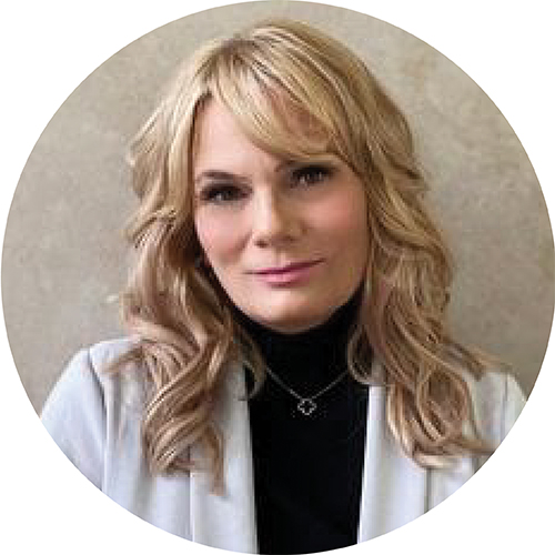 Portrait framed in a circle. Subject is looking at the camera and can be seen from the shoulders up. They are wearing a grey jacket with a black turtle neck, with blonde hair down past their shoulders, standing against a tan backdrop.