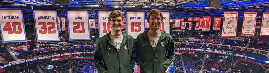 Twins pose together with Pistons Jersey's behind them at a sports arena.