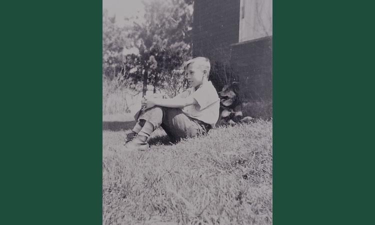 Larry Leinweber as a child sitting on the ground outside his rural home