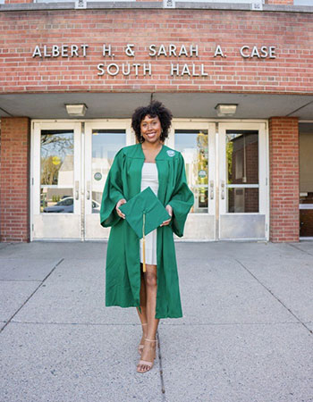 A student in a graduation gown stands in front of a brick building.