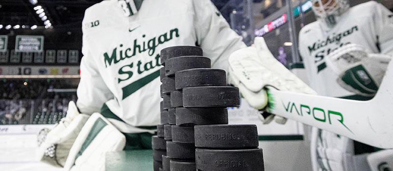 an artistically cropped photo shows a stack of hockey pucks balanced on the boards as two hockey players in goalie gear make their way out onto the ice
