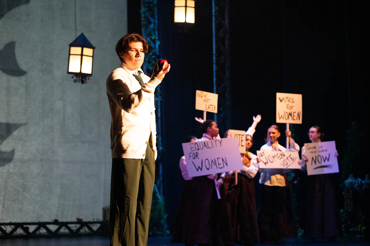 A theatre performer stands in the foreground holding an apple while a group of women performers stands behind him hold suffrage signs.