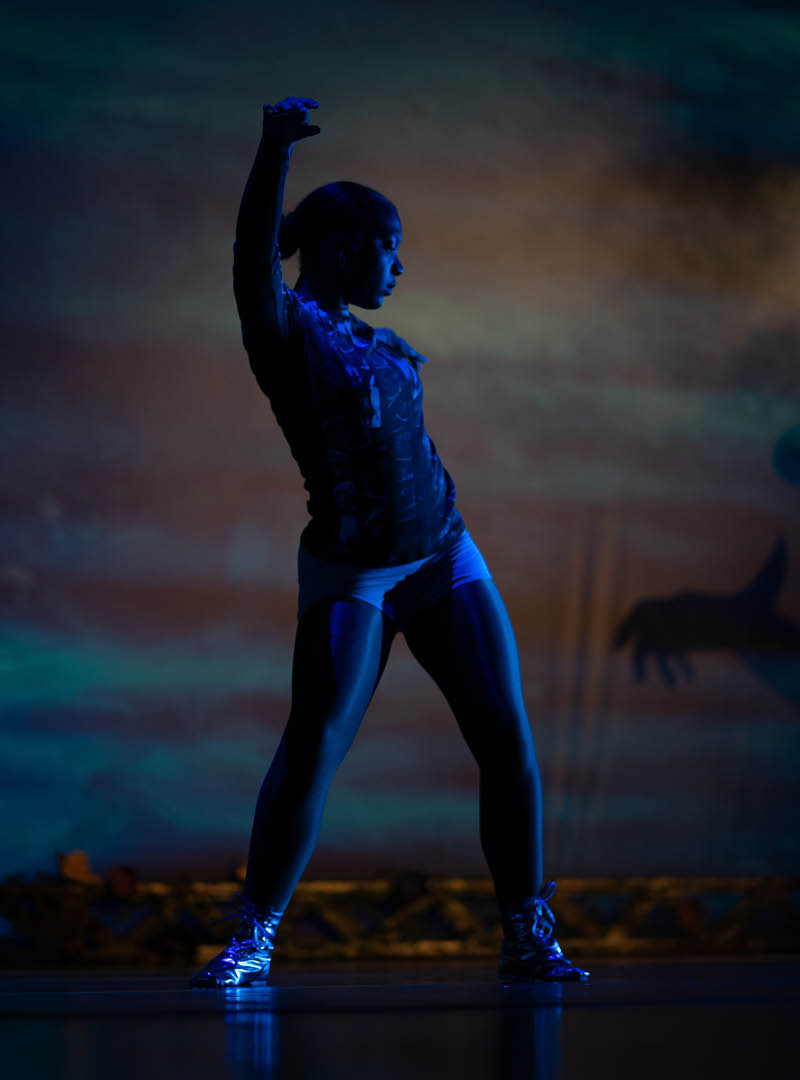 A performer dances in low lighting on stage during a theatre production.
