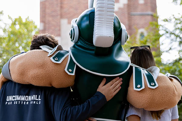Students pose with Sparty in front of Beaumont Tower on the Michigan State University campus, with one student wearing a jacket displaying the campaign message, “Uncommon Will. Far Better World.”