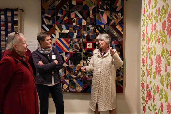 MSU alumna Christine Force leads a tour group standing in front of three mutli-colored quilts hanging on the walls at the Maine Historical Society.