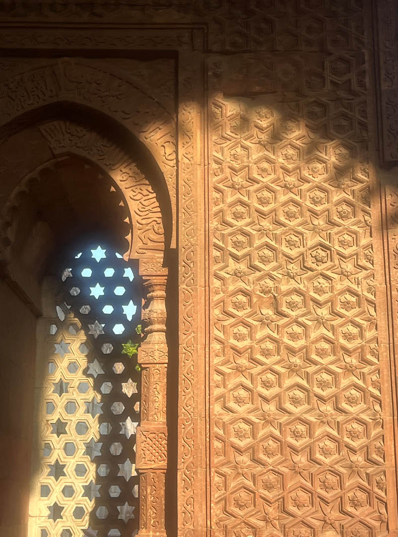 Decorative star and pentagon shapes cross through sun and shadow on a red sandstone window and wall with inlaid white marble.