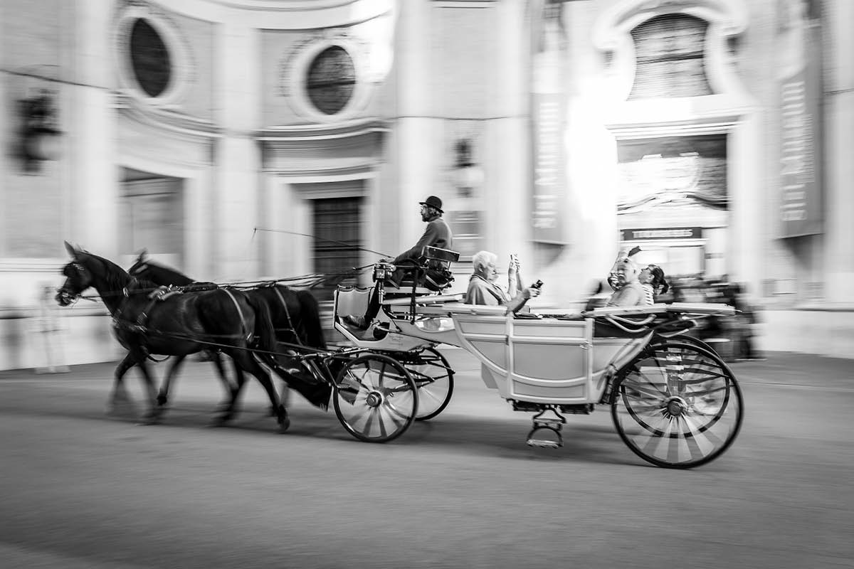 A black and white photo of a horse-drawn carriage as is rolls down the street.