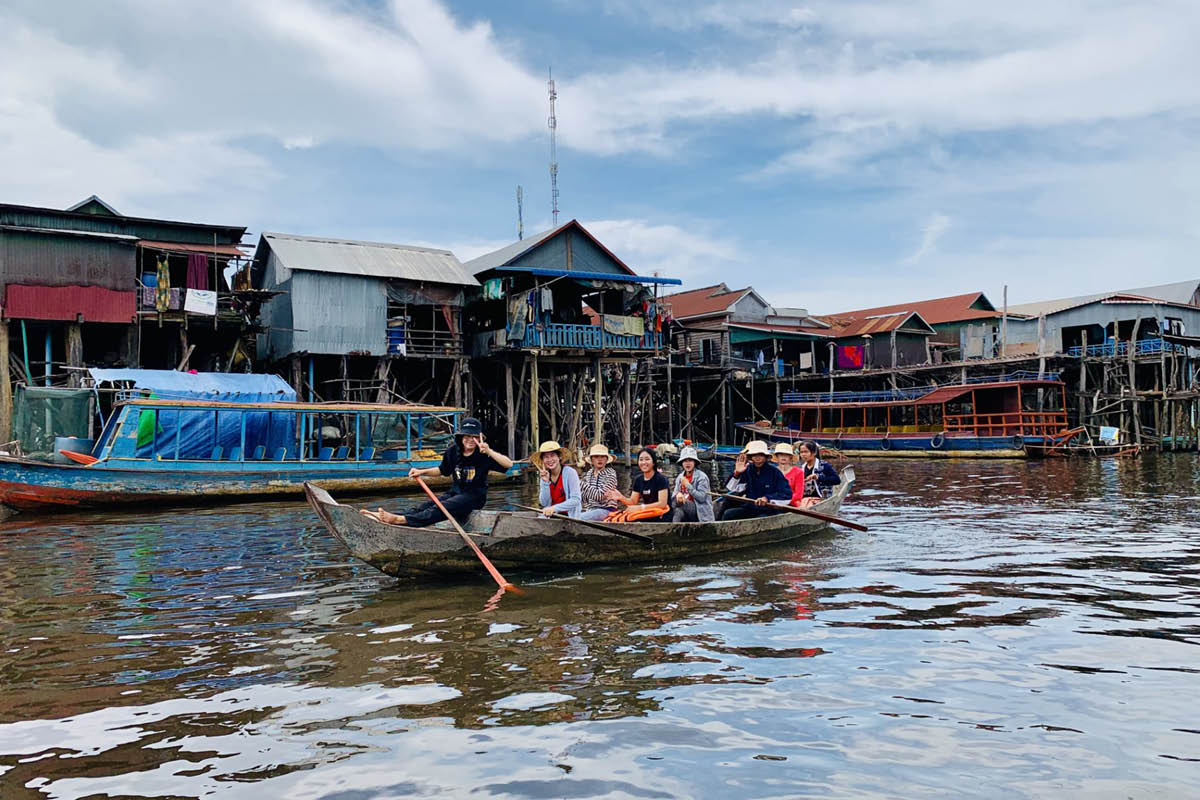 A group of people ride in a long wooden boat through a waterside village with stilted homes and docked boats. One person paddles at the front while others smile toward the camera beneath a cloudy blue sky.