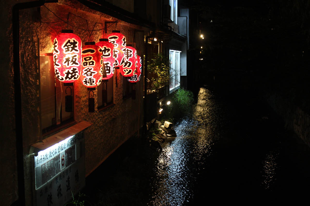 A row of oriental lanterns with Chinese characters hang off the side of a dark building over a river a night.