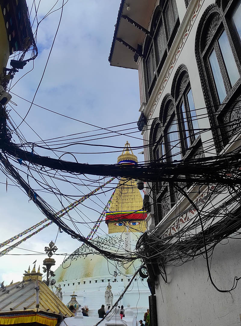 Utility wires drape across an alley in the foreground with the top of the Boudhanath stupa in Kathmandu in the background.