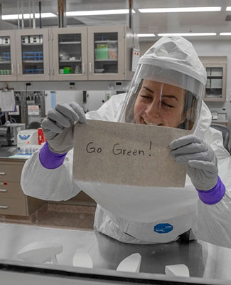 A scientist in full PPE holds up a handwritten banner that says Go Green through the window of a lab at the MSU VDL.