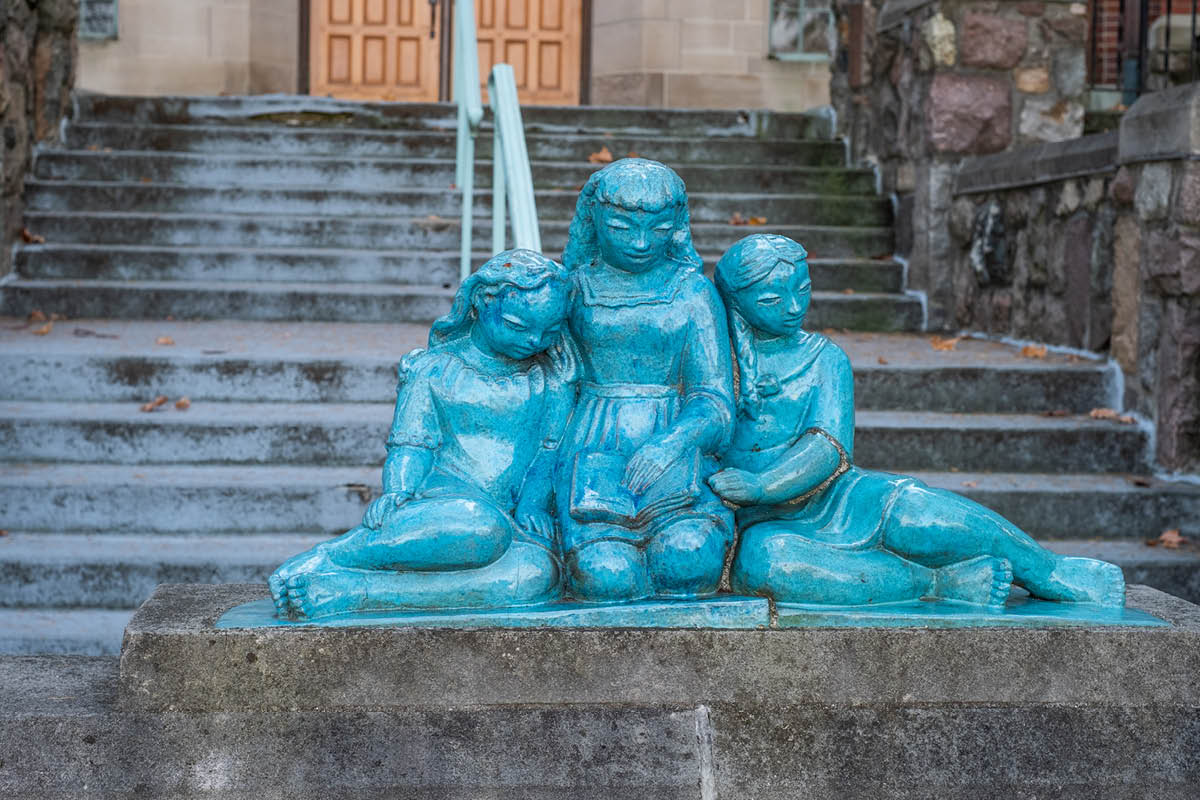A light blue Pewabic Pottery sculpture of three young girls sitting together called 'Children Reading and Fish Waterspout' located at the Williams Hall north entrance.