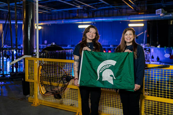 MSU alums Dana MacCallumMhor and Lauren Grauer stand together holding a Michigan State Spartan flag in front of yellow railings surrounding the top of a large aquarium tank.