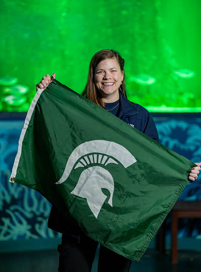 Alumna Lauren Grauer holds up a green Michigan State flag with a white Spartan logo, while in the background, fish swim past the window of an aquarium tank lit up with green lights.
