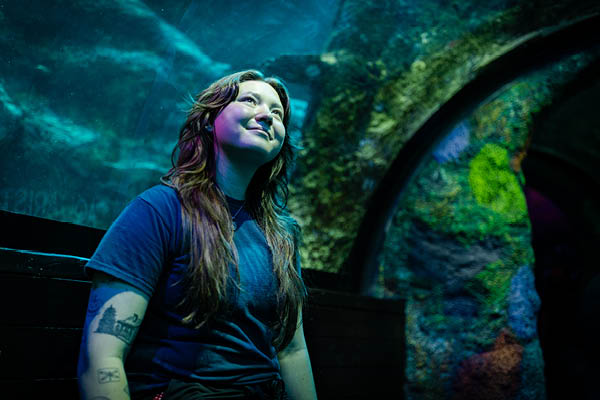 MSU alumna Dana MacCallumMhor stands inside a Sea Life Aquarium tunnel, looking upward with a soft smile as blue-green light from the surrounding water illuminates her face; fish tanks and curved glass arches frame the scene.