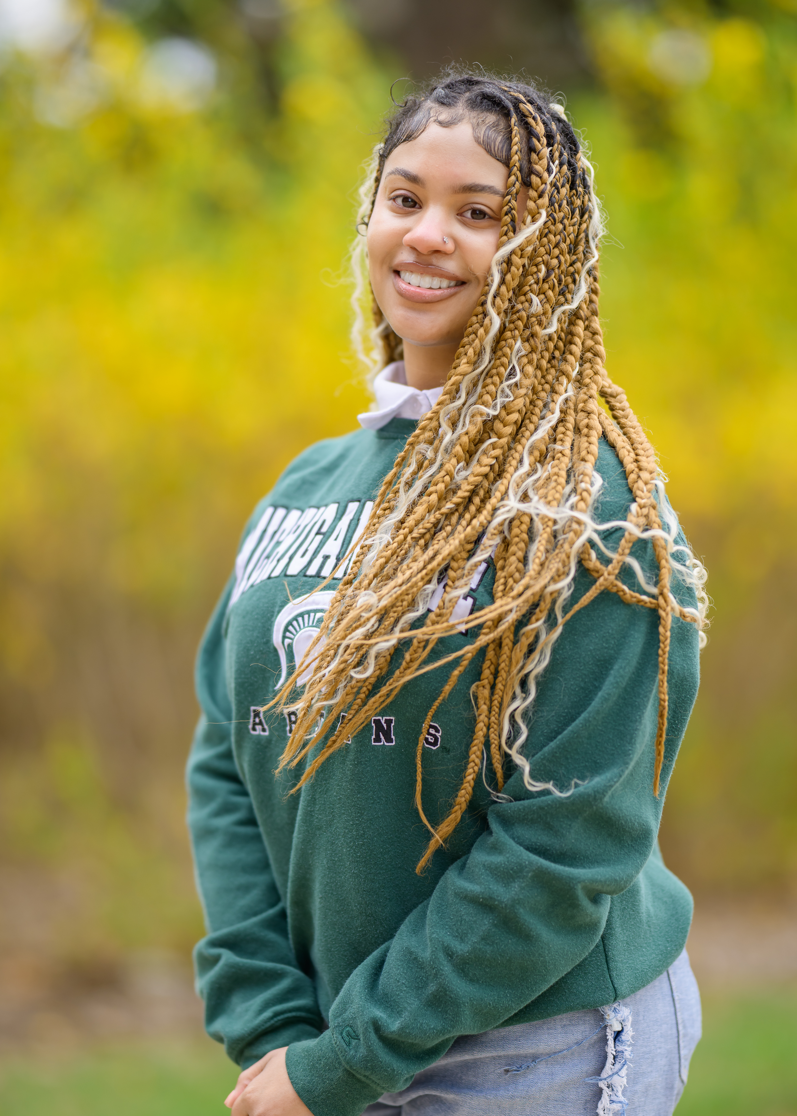 Jaelynn Smith, a member of the 2025 MSU Homecoming Court, smiles brightly while standing outdoors in front of vibrant yellow foliage. She wears a green Michigan State Spartans sweatshirt and ripped jeans, with her long, golden and blonde braids styled down over one shoulder.