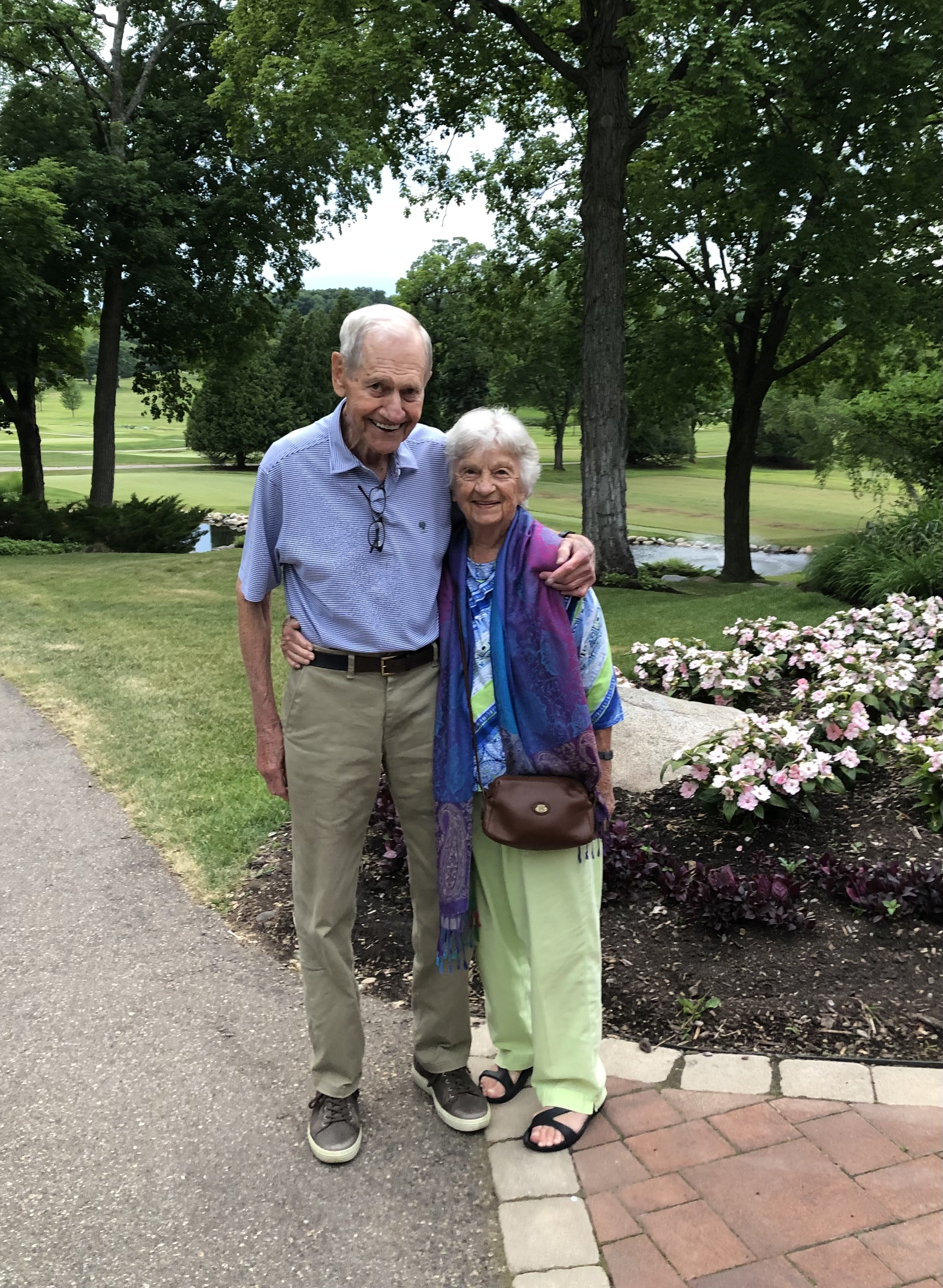 Hal and Peggy Bernthal smiling together on a garden path surrounded by trees and flowers.
