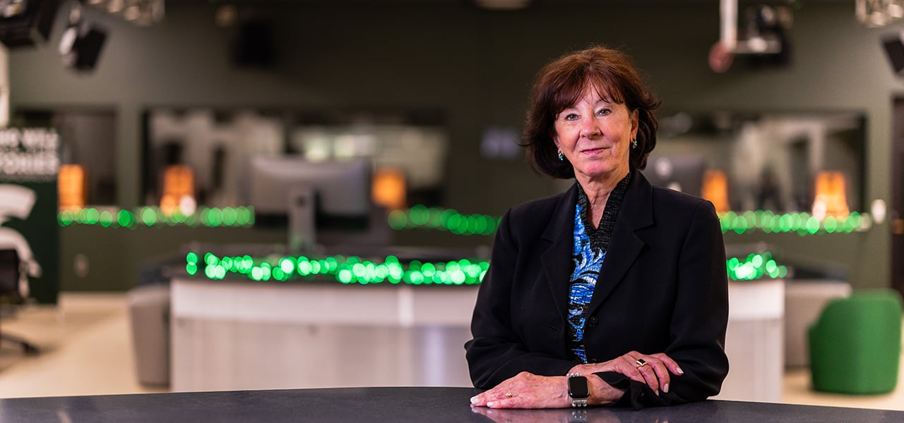 Branstetter wearing a dark blazer standing at a table in a modern studio with green lights in the background.