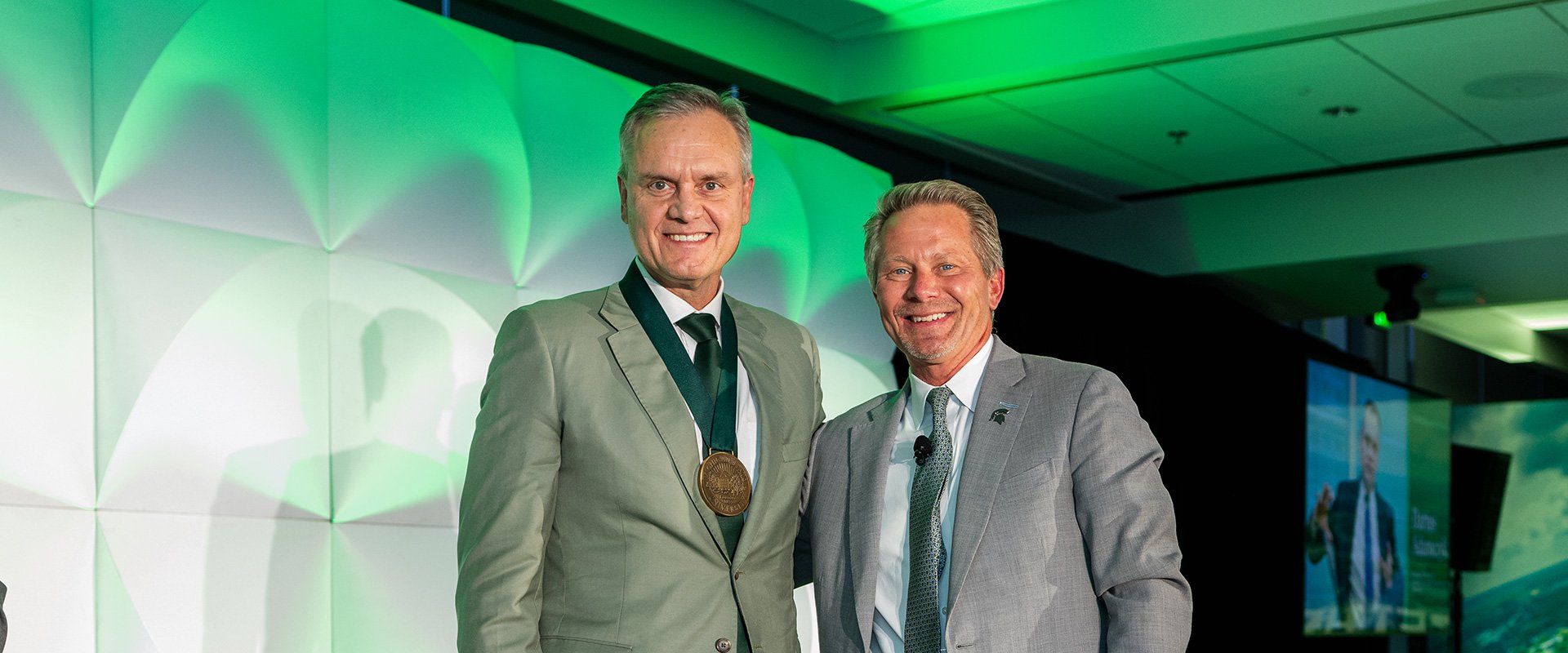 Adamczyk and President Guskiewicz in suits standing on a stage with green lighting, one wearing a medal, near a table with awards and a screen displaying 'TOGETHER'.