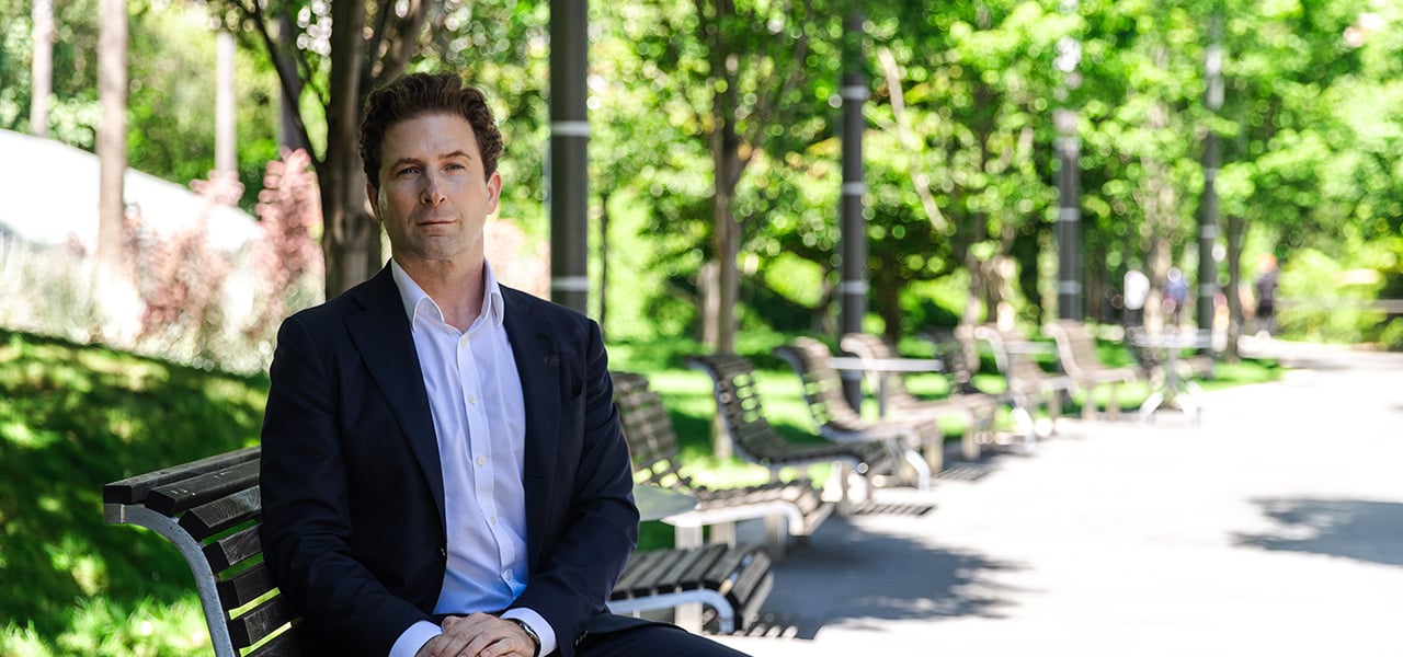Dean Fealk wearing a dark suit jacket and white dress shirt is seated on a curved metal bench along a tree-lined pathway in a park-like setting.