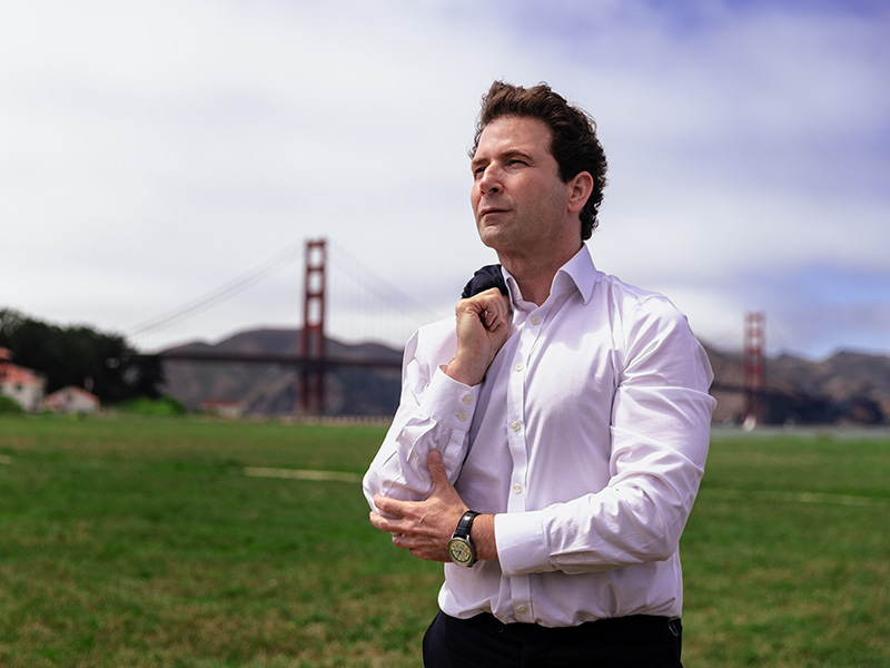 Dean Fealk wearing a white dress shirt and dark pants stands on a grassy field holding a jacket over one shoulder. In the background, the Golden Gate Bridge spans across the scene with hills and buildings visible under a partly cloudy sky.